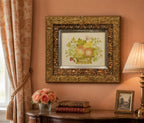 Decorative painting in an ornate gold frame on a wall with a lamp and books on a table.