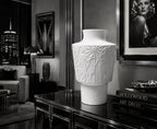 White decorative vase on a table with a cityscape and portrait in the background