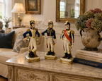 Three decorative figurines of soldiers on a marble table with books and a vase in the background.
